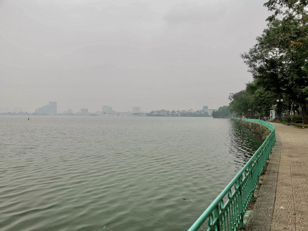 A view across the vast surface of West Lake in Hanoi on a hazy day. A curved, green metal railing follows the paved pathway along the shoreline, with the city's hazy skyline visible in the distance across the water.