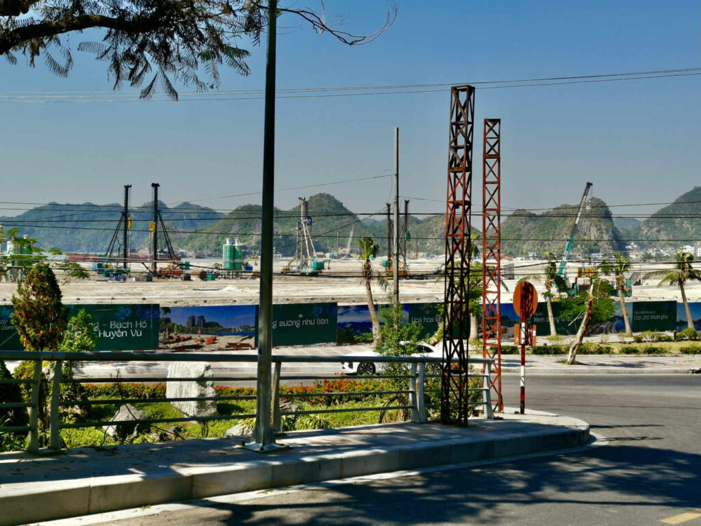 Construction site on Cat Ba Island with cranes, drilling rigs, and cleared land, showing large-scale development against a backdrop of green limestone hills.