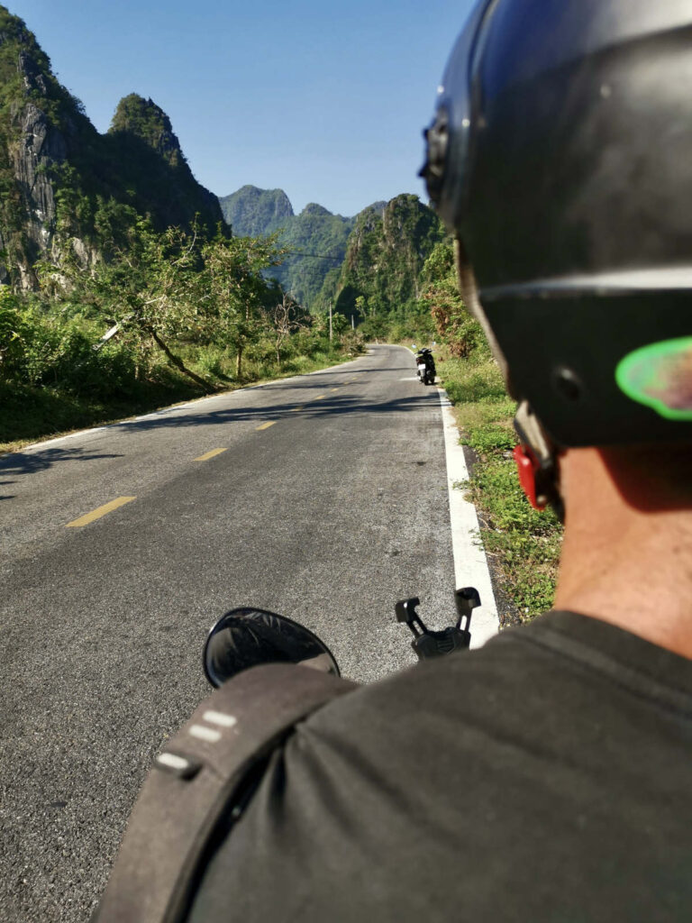 A person on a scooter, wearing a black helmet and t-shirt, looks down a winding road on Cat Ba Island, Vietnam. The road, with a yellow dividing line, is flanked by lush green foliage and towering limestone mountains under a clear blue sky. Another scooter is visible in the distance.