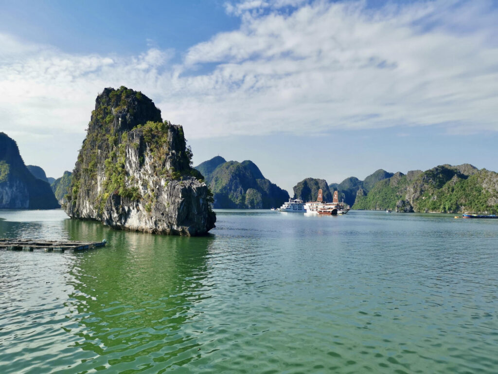 A wide shot of Halong Bay, Vietnam, featuring multiple limestone karst islands jutting out of the emerald green water under a blue sky with scattered clouds. In the foreground, a large, prominent island is covered in lush green vegetation. In the middle ground, two cruise ships are visible, one a modern white vessel and the other a traditional junk boat with brown sails. A wooden floating structure is in the left foreground.