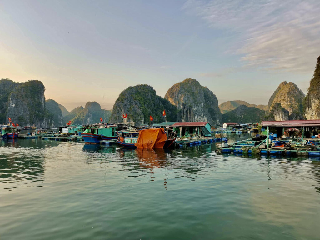 A collection of small, colorful houses built on floating platforms in a sheltered cove in Ha Long Bay, Vietnam. Several small fishing boats are moored in front of the houses, and the iconic, lush limestone mountains surround the village under a partly cloudy sky.