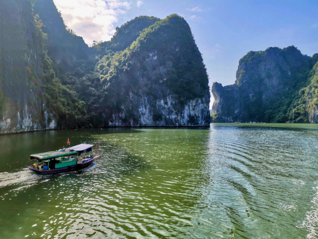 A boat with a green roof and flags sails across the emerald green water of Ha Long Bay, leaving a wake behind it. Towering, tree-covered limestone karsts with sheer cliffs rise dramatically from the water under a blue sky with some wispy clouds. The sun shines brightly on the water.