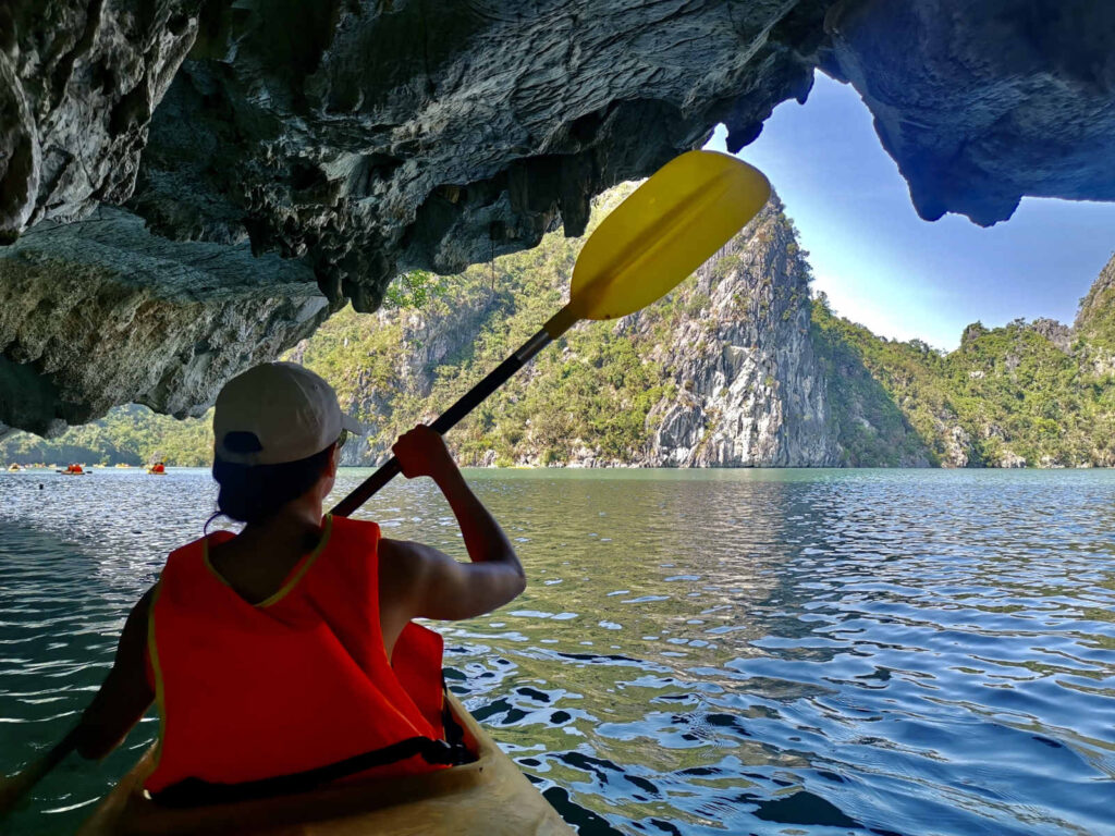A person wearing an orange life vest and a white hat is seen from behind, paddling a yellow kayak out of a dark, rocky sea cave. The kayak is on a calm, turquoise body of water surrounded by steep, jungle-covered limestone cliffs under a bright blue sky.