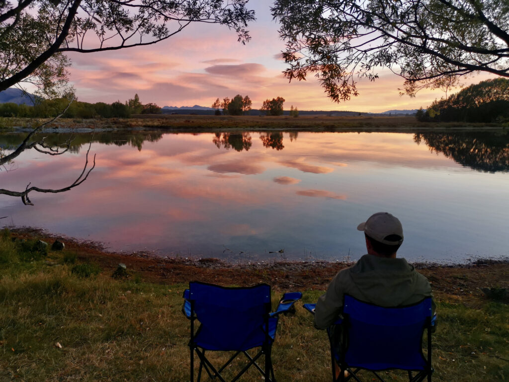 Two blue camping chairs sit on a grassy bank overlooking the calm, reflective water of a lake at sunrise, with a person sitting in one of the chairs and a stunning pink and orange sky reflected in the water.