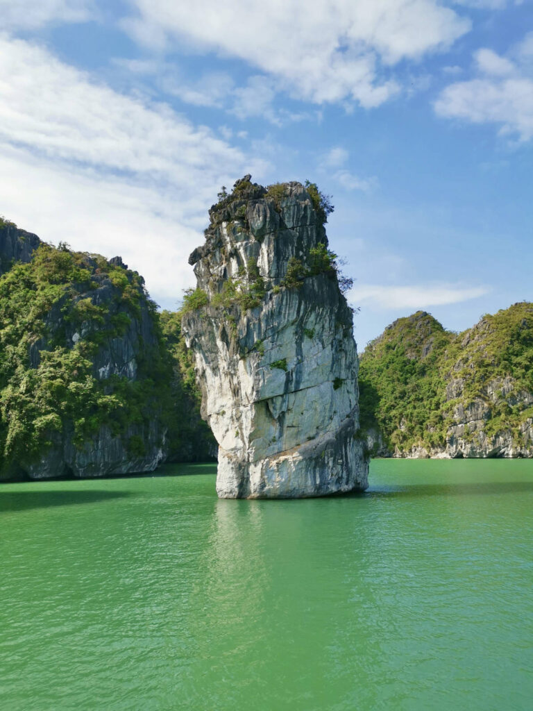 A large, distinctive, and unusually shaped limestone karst rock formation rises dramatically from the calm, emerald-green water of a bay. The rock is mostly light gray with some white streaking and is topped with a cluster of green trees. Smaller, tree-covered islands and a few distant boats are visible in the background under a blue sky with scattered clouds.