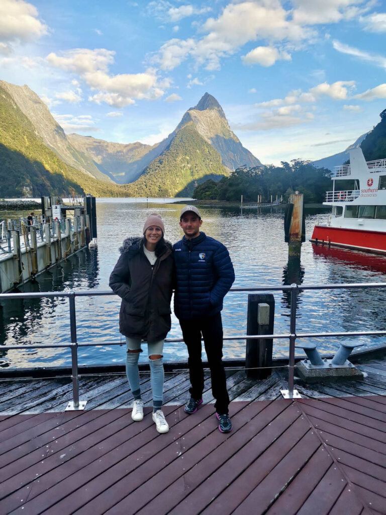 Katharina and Allan smile for a photo on a wooden pier in Milford Sound, New Zealand, with the iconic, cone-shaped Mitre Peak and the calm waters of the fiord in the background.