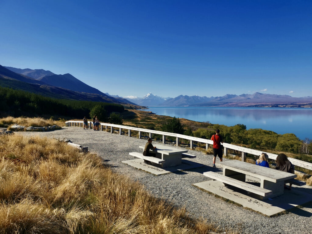 People are seated at picnic tables and walking along a lookout point, with a stunning view of the turquoise waters of Lake Pukaki and the snow-capped mountains of Aoraki/Mount Cook National Park in the distance under a clear blue sky.