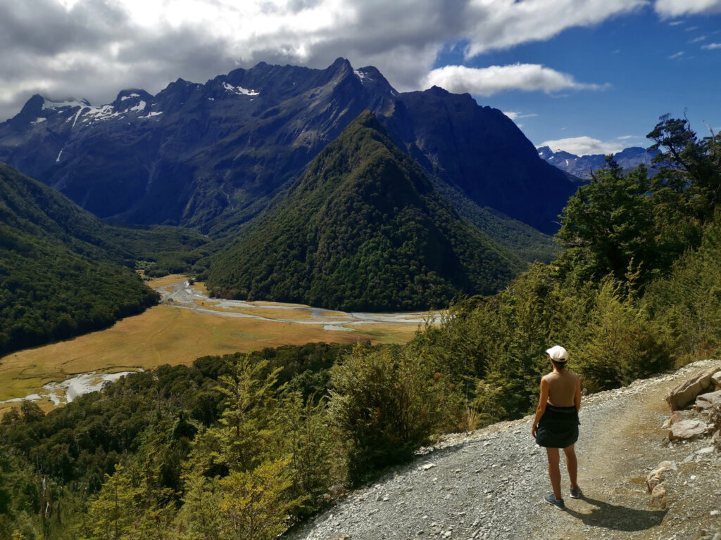 Katharina wearing a hat and shorts stands on a gravel path, looking out at a stunning mountain valley with a winding river and a large, green-covered hill in the center, with towering, snow-dusted mountains in the distance under a partly cloudy sky.