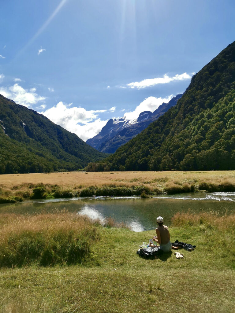 Katharina sits on the grassy bank of a calm river in a glaciated valley on the Routeburn Track, looking out at the stunning mountain scenery with snow-capped peaks in the distance under a sunny sky.
