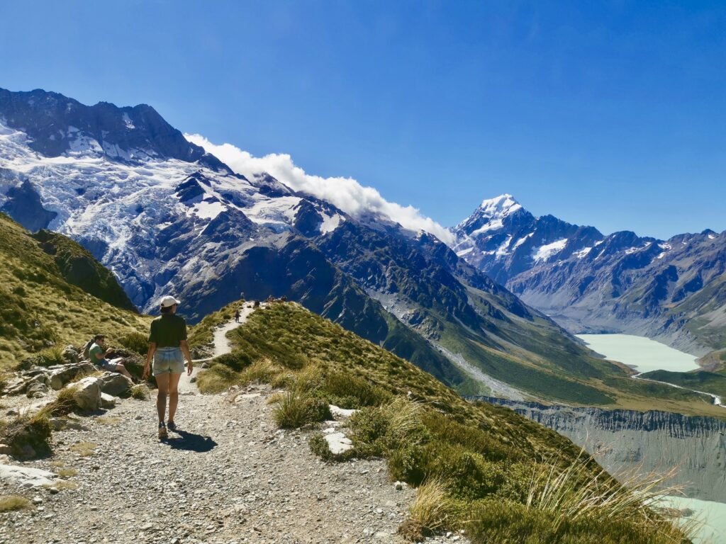 Katharina walking along a gravel path on the Sealy Tarns track, with a view of a mount cook and a glacial lake below.