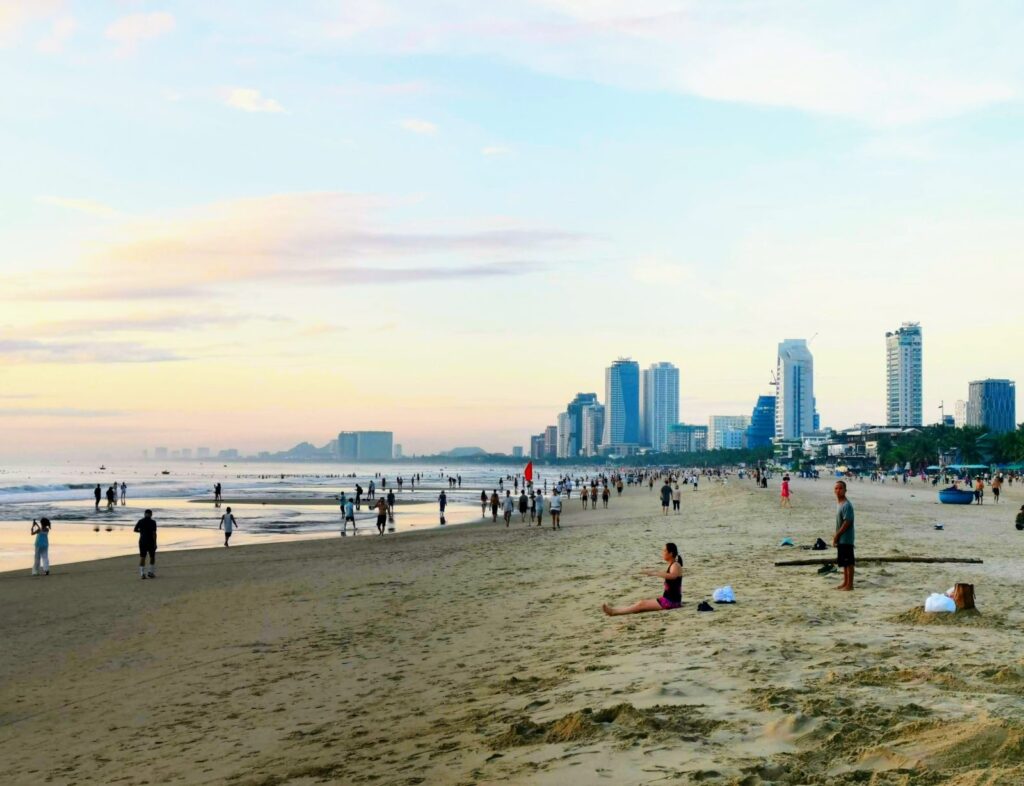 Wide Da Nang beach at sunrise (6 AM) showing hundreds of locals exercising, walking, and enjoying the sand in front of the city skyline.