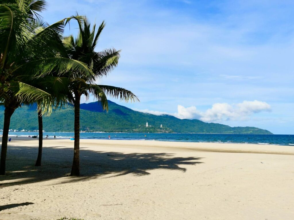 Wide expanse of Da Nang beach in Vietnam, showing palm trees casting shadows on the white sand with Monkey Mountain (Son Tra Peninsula) and the Lady Buddha statue visible in the background.