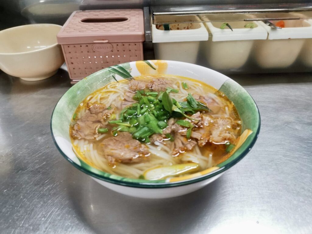 Close-up of a bowl of Bún Bò Huế, a spicy Vietnamese beef noodle soup topped with fresh green onions and herbs, photographed in Da Nang.