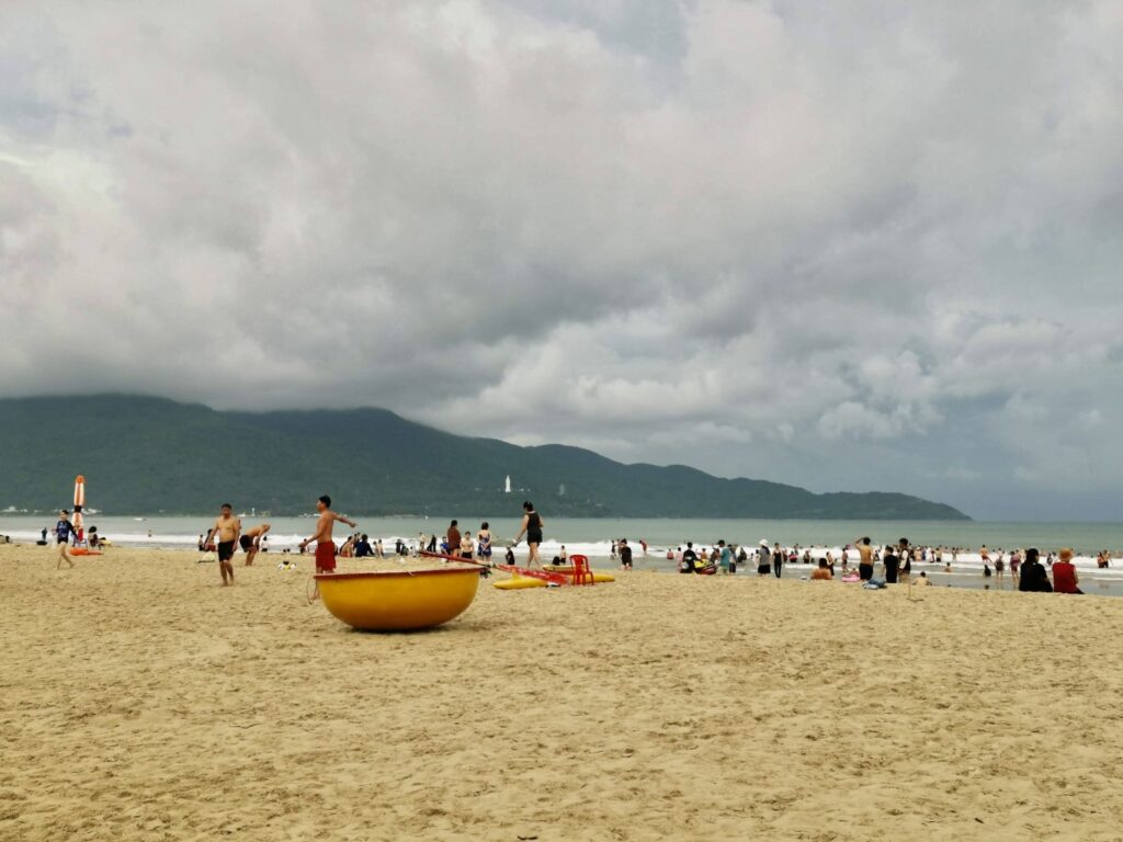 Overcast and cloudy day at Da Nang's My Khe Beach during the rainy season, with a few people scattered on the wide, golden sand and mountains visible in the distance.