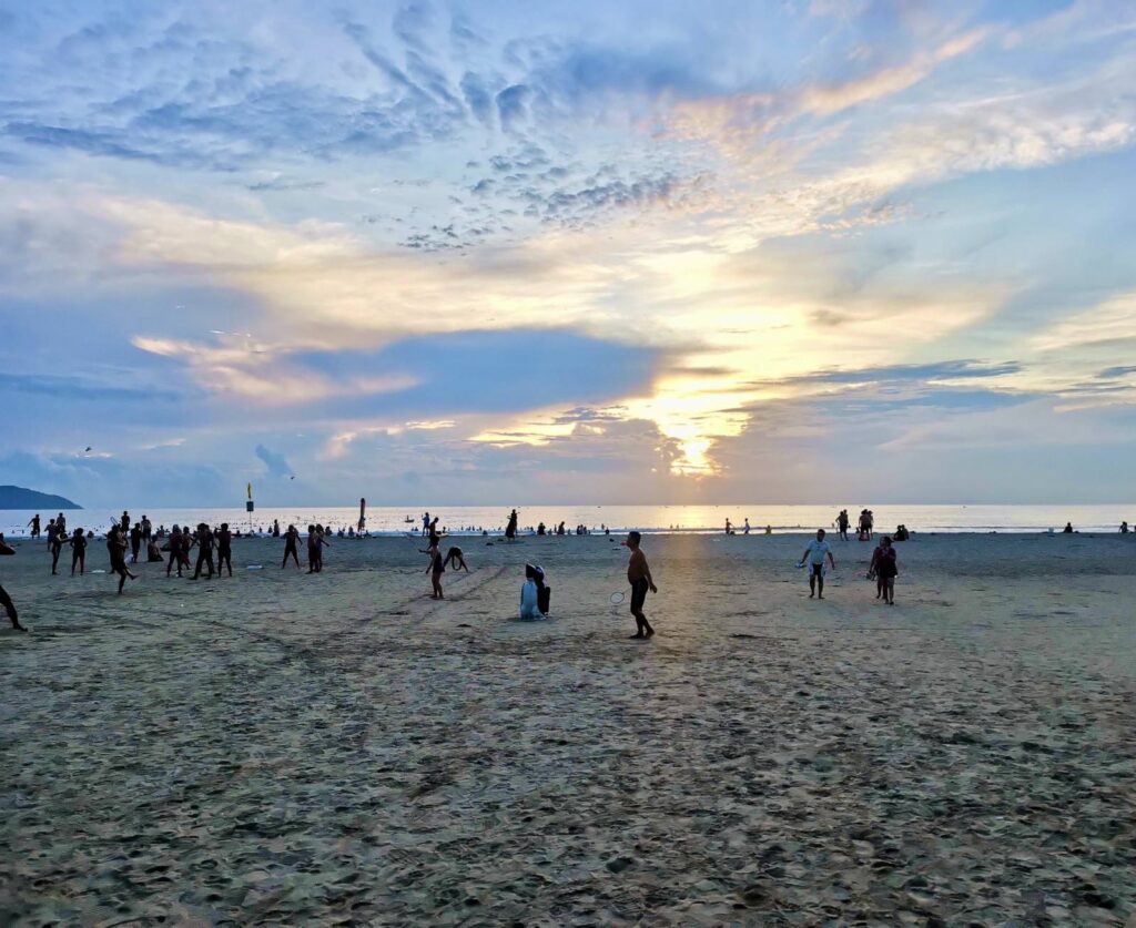 Da Nang's My Khe Beach at sunrise, crowded with hundreds of locals swimming, exercising, and playing sports, showcasing the city's vibrant early morning community atmosphere.
