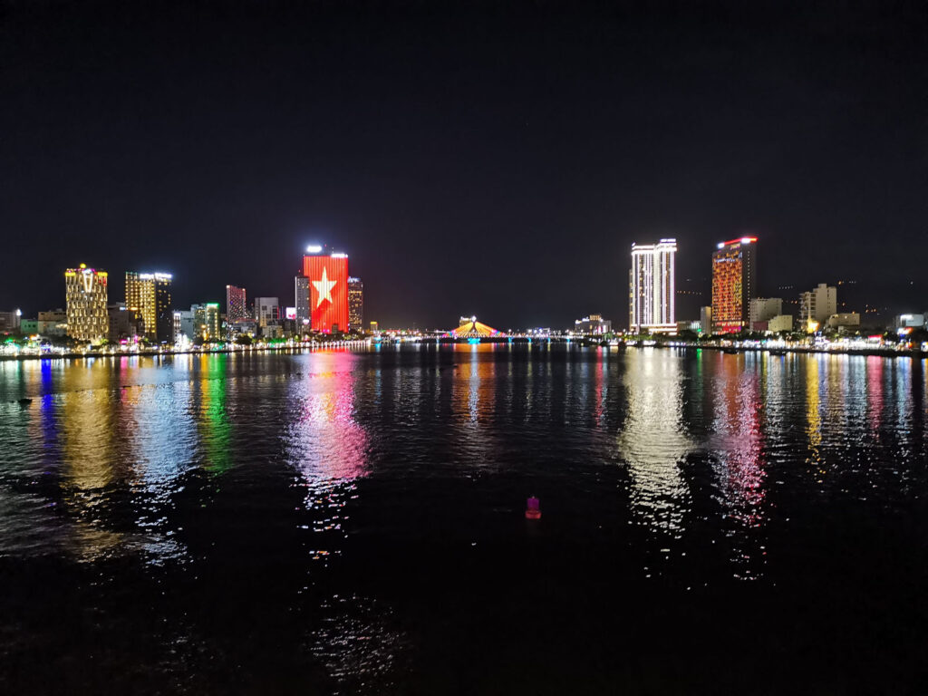 Da Nang city skyline at night, showing brightly illuminated high-rise buildings and the reflection of the lights in the dark water of the Han River.
