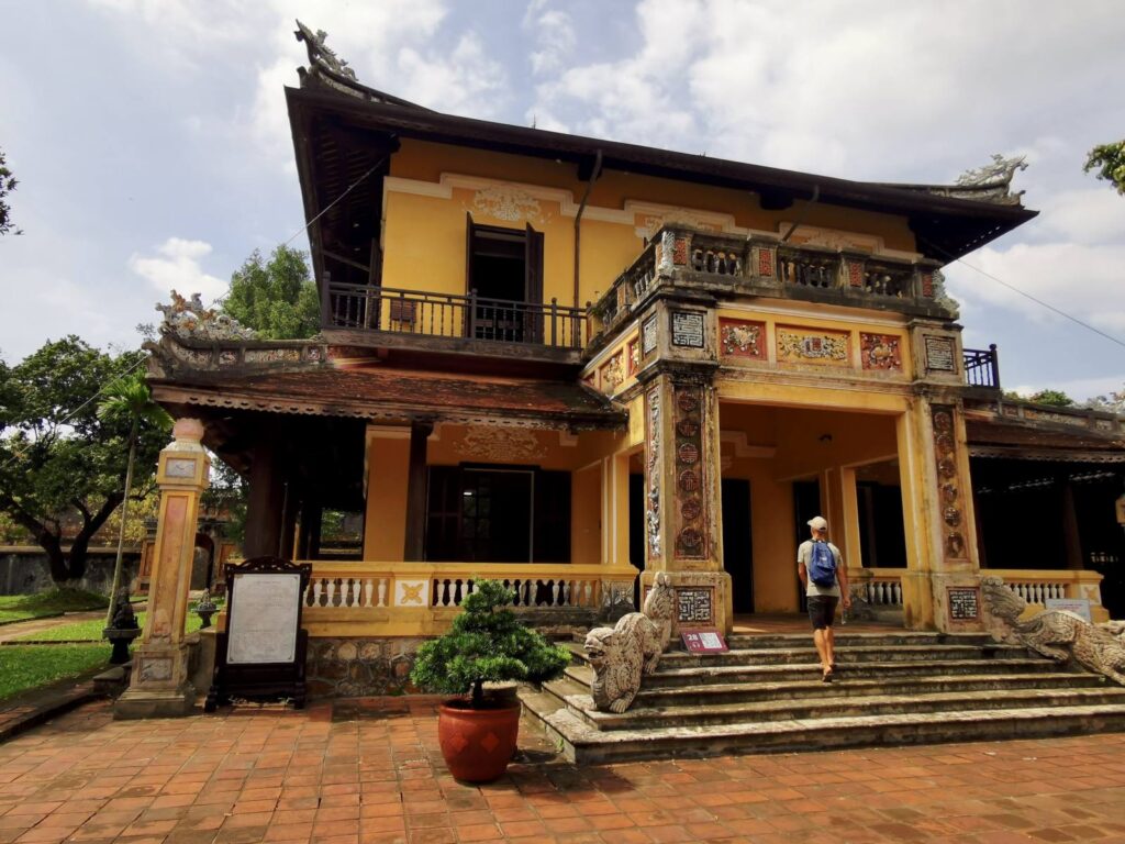A traveler walking toward the Lau Khuong Ninh pavilion in the Dien Tho Palace residence, a yellow two-story building with a traditional balcony and stone dragon steps in Hue.