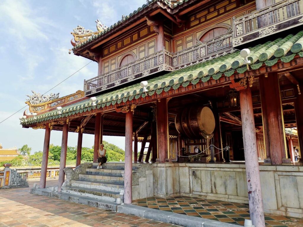Katharina sitting on the steps of the ornate, multi-story Meridian Gate in Hue, Vietnam, featuring green tiled roofs and a large traditional drum in the background.
