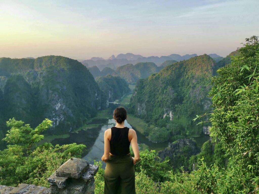 Katharina standing at the Hang Mua viewpoint in Ninh Binh, looking out over a winding river valley surrounded by steep limestone karst mountains at sunset.