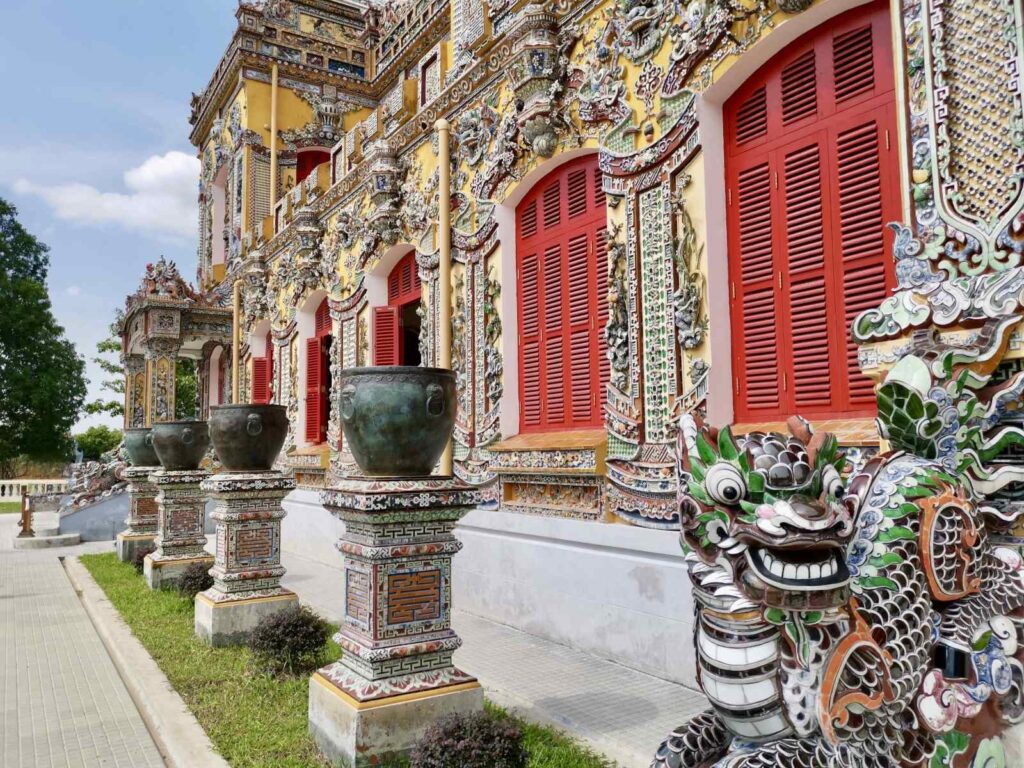 A close-up of the detailed architecture at Kien Trung Palace in Hue, featuring a colorful ceramic dragon statue, large bronze urns, and intricate porcelain mosaic wall art.