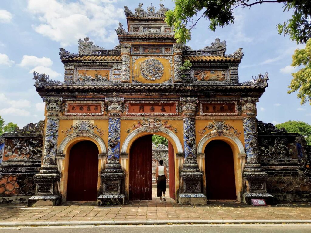 A traveler walking through a massive, ornate yellow and blue ceramic gate at the Imperial City (Hoàng Thành) in Hue, Vietnam.