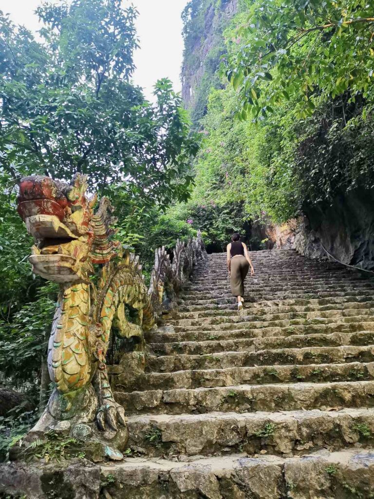 Katharina in green pants climbing the ancient-style stone stairs at Mua Cave, Ninh Binh, next to a large decorative dragon sculpture and lush green cliffs.