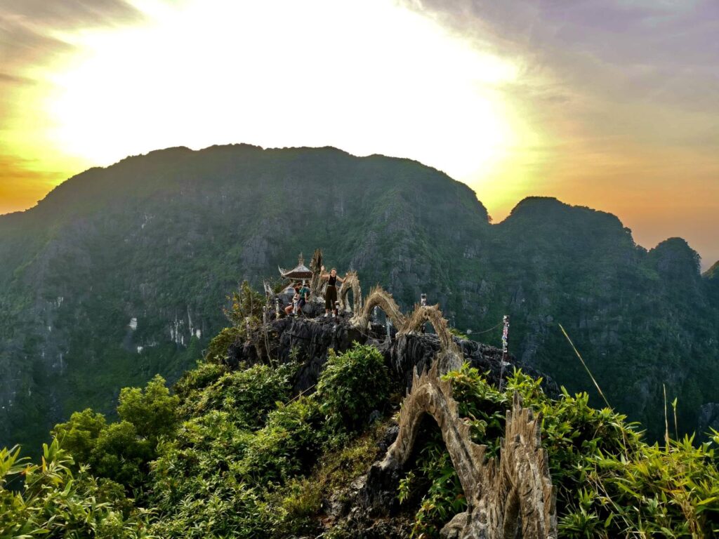 A traveler standing next to the ornate stone dragon sculpture on the summit of Hang Mua at sunrise, with a bright sunburst over the limestone mountains.