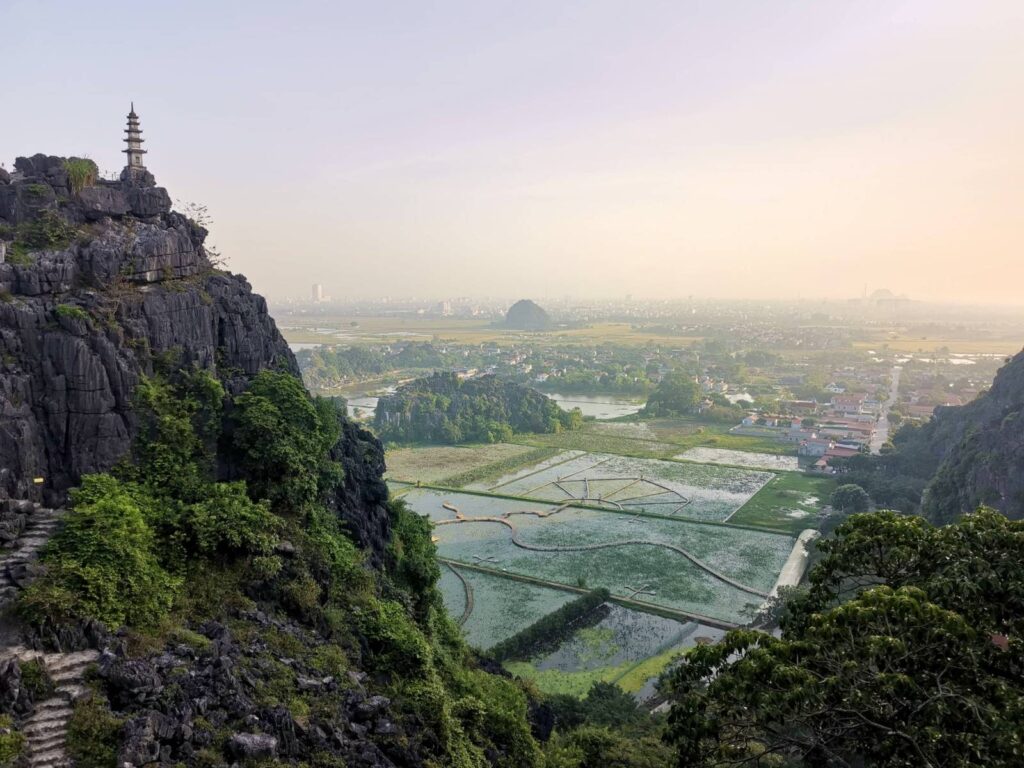 A wide landscape view from Hang Mua looking across at the pagoda perched on a limestone peak with the heart-shaped lotus pond boardwalk in the valley below.