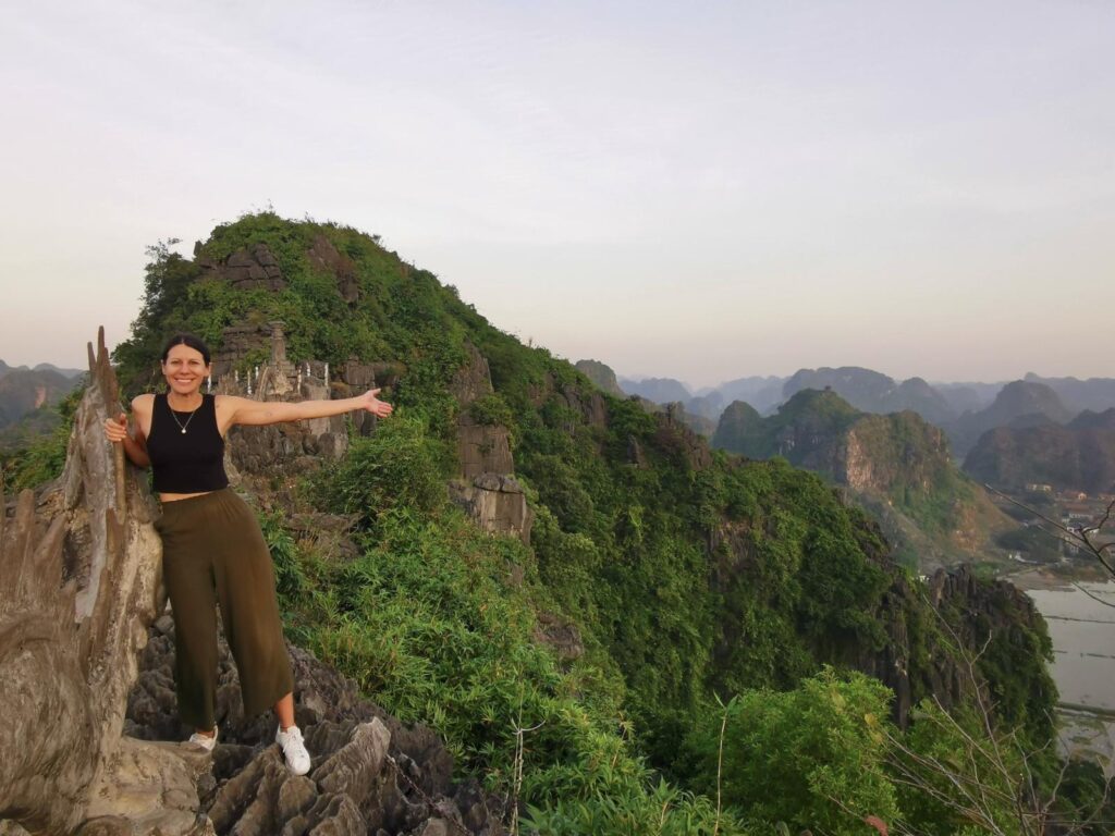 Katharina smiling and gesturing toward the jagged limestone peaks at the top of Hang Mua in Ninh Binh, standing next to a stone dragon carving with a panoramic valley view in the background.