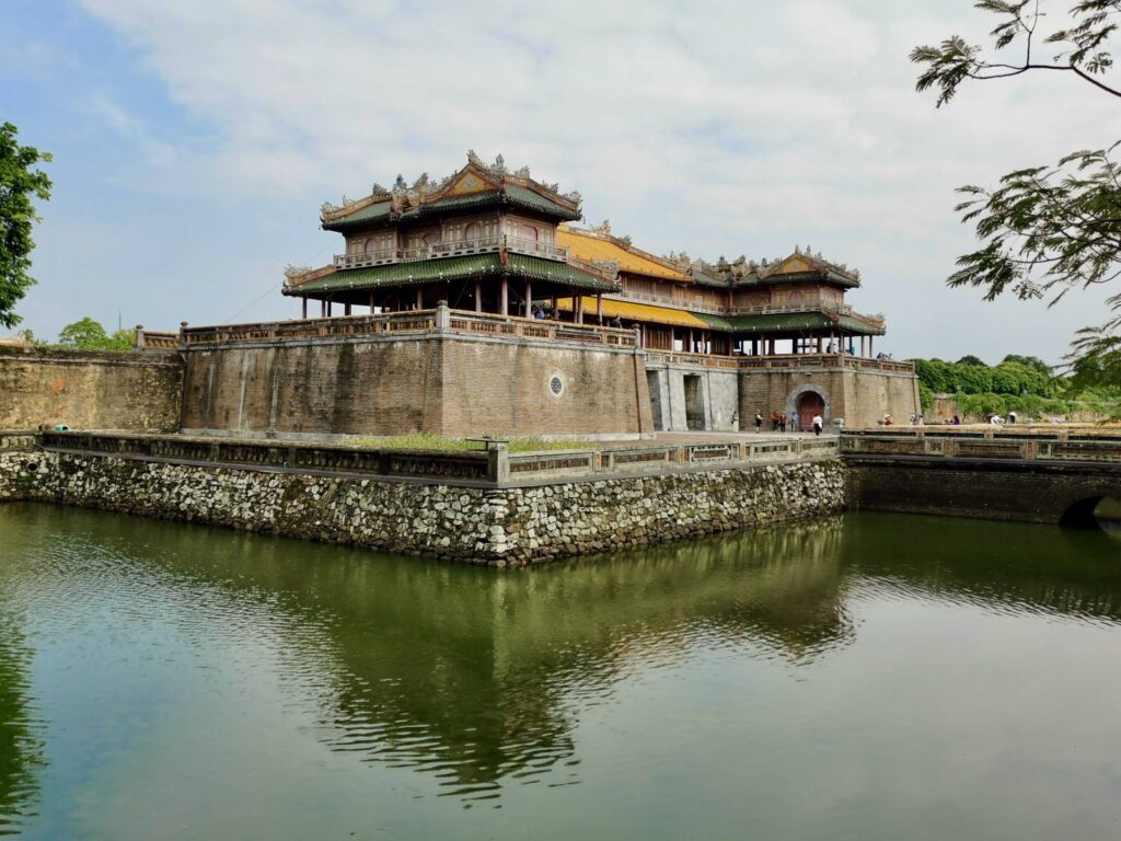 A scenic view of the Ngan Gate in Hue, Vietnam, featuring a stone bridge crossing a calm moat that leads into the fortified three-arched gateway of the Imperial Citadel.