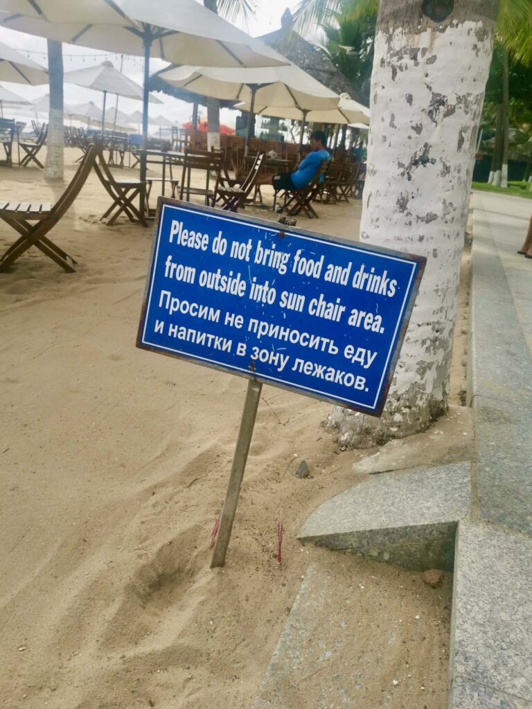 Sign on Nha Trang beach written in both English and Russian, warning tourists not to bring outside food into the sun chair area, highlighting the large Russian community presence.
