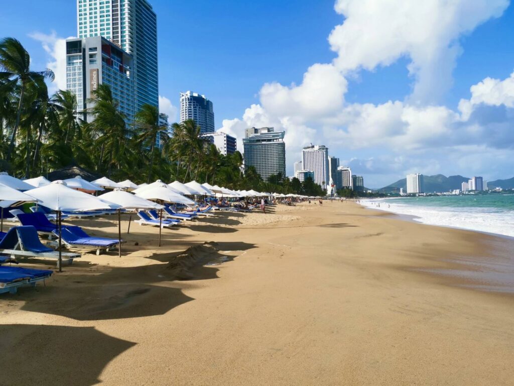 Steep sandy Nha Trang beach lined with white umbrellas and sun loungers, showing high-rise hotels and the urban skyline directly behind the beach.