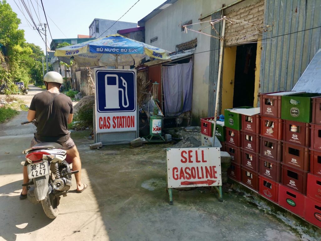 A man on a motor scooter stopping at a local roadside fuel station in Vietnam with signs saying 'Cas Station' and 'Sell Gasoline' near Ninh Binh.
