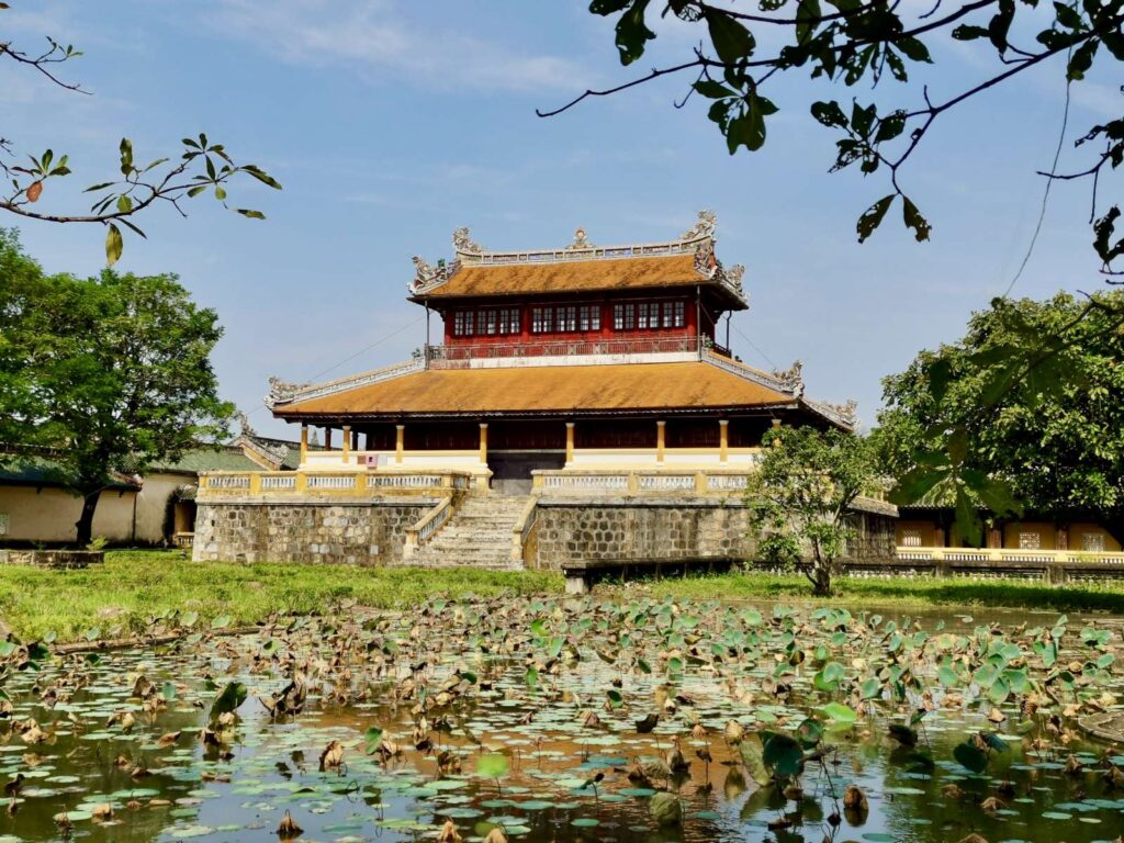 A wide shot of the two-story Thai Binh Reading Pavilion in Hue, featuring a bright yellow tiled roof and red upper walls, standing behind a large pond filled with green lotus leaves.