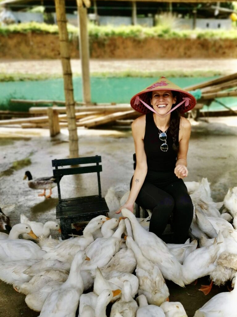Katharina smiling wearing a traditional Vietnamese conical hat (Non La) sitting on a stool and surrounded by a large flock of white ducks at The Duck Stop in Phong Nha.