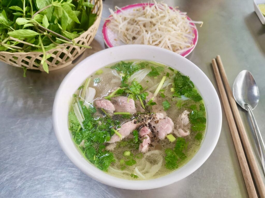 A top-down view of a steaming bowl of beef pho at Pho Kim Huyen in Hue. The bowl is filled with clear broth, rice noodles, tender beef slices, and fresh cilantro. It is served on a metal table alongside a basket of fresh green basil, a plate of bean sprouts, and a pair of wooden chopsticks.