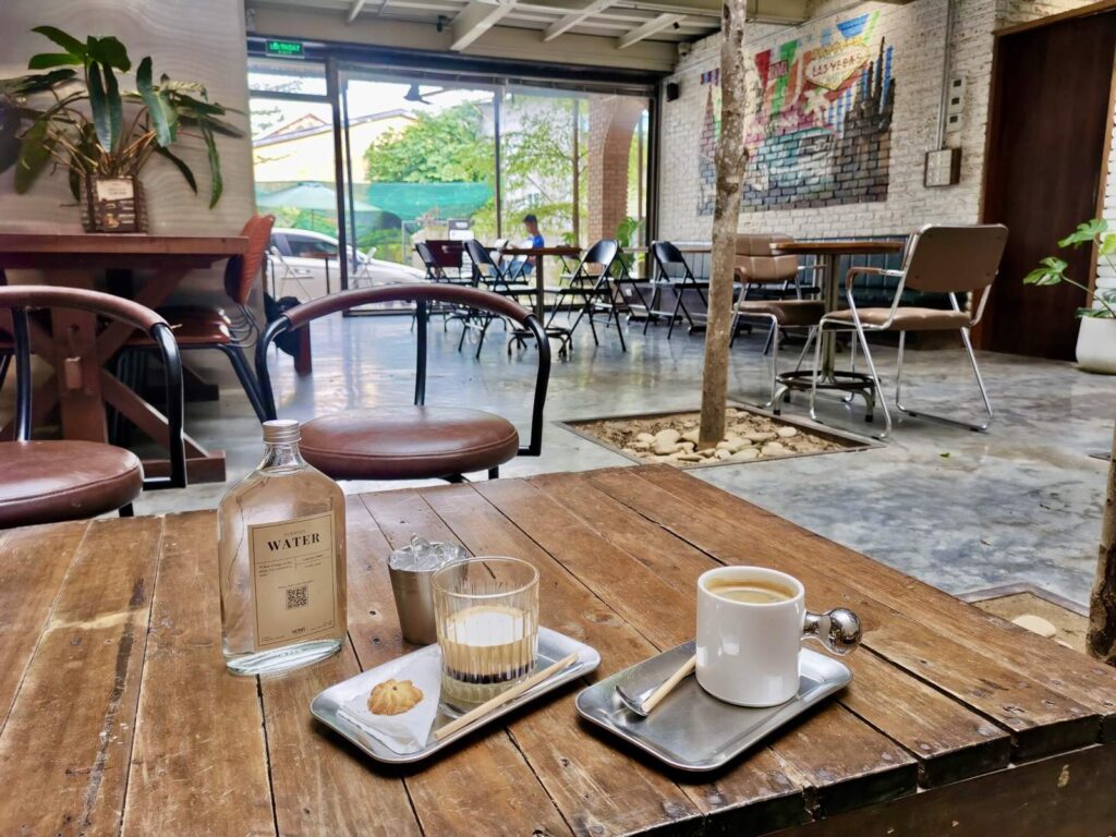 A modern, industrial-style cafe interior at Venus iCafe in Hue, featuring a wooden table in the foreground with a glass of water, a cookie, and a cup of coffee. The background shows a bright, open seating area with large glass windows, indoor trees, and a brick wall with a colorful mural.