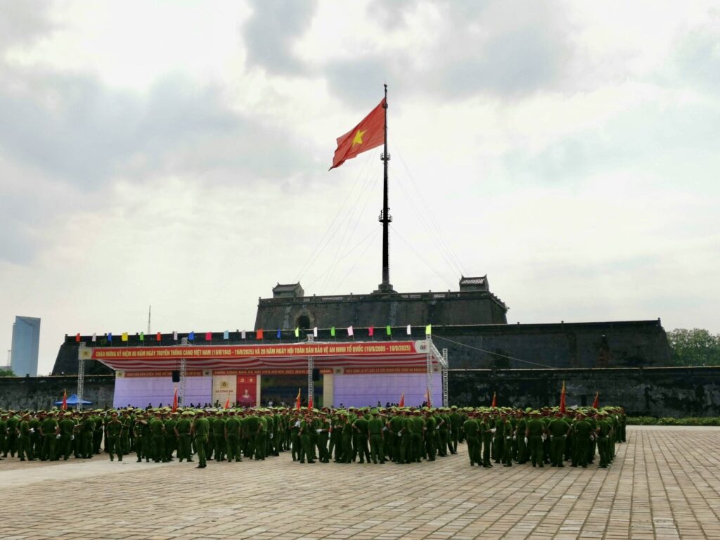 large group of Vietnamese officers in green uniforms gathered in a paved plaza in front of the massive stone Flag Tower flying the Vietnamese national flag in Hue.