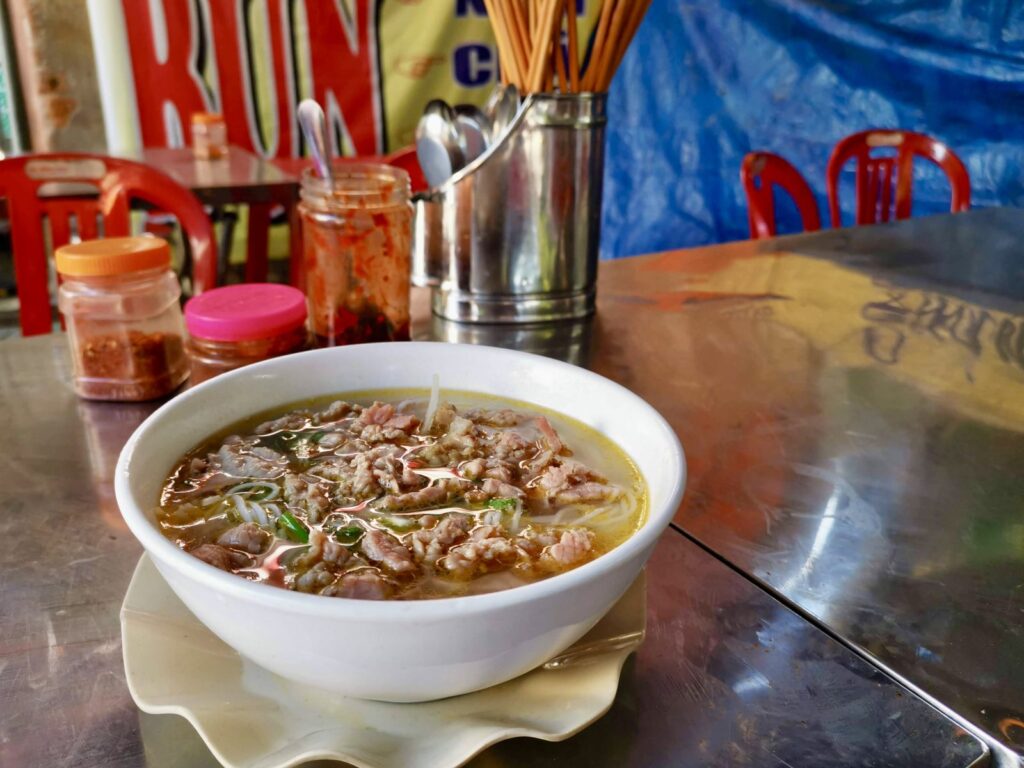 A bowl of bun bo Hue with rich broth, sliced beef, and noodles sits on a metal table at a local eatery in Hoi An. Condiment jars and chopsticks in the background show a casual Vietnamese street food setting.