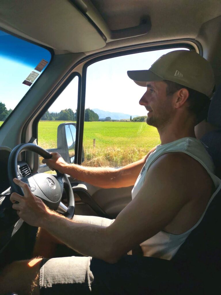 Allan driving a campervan past bright green fields and distant mountains on a sunny day on New Zealand's South Island.