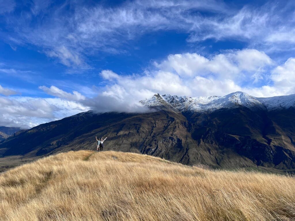 Allan and Katharina standing on a golden hilltop facing snow-capped mountains at the end of the Diamond Lake Hike near Wanaka.