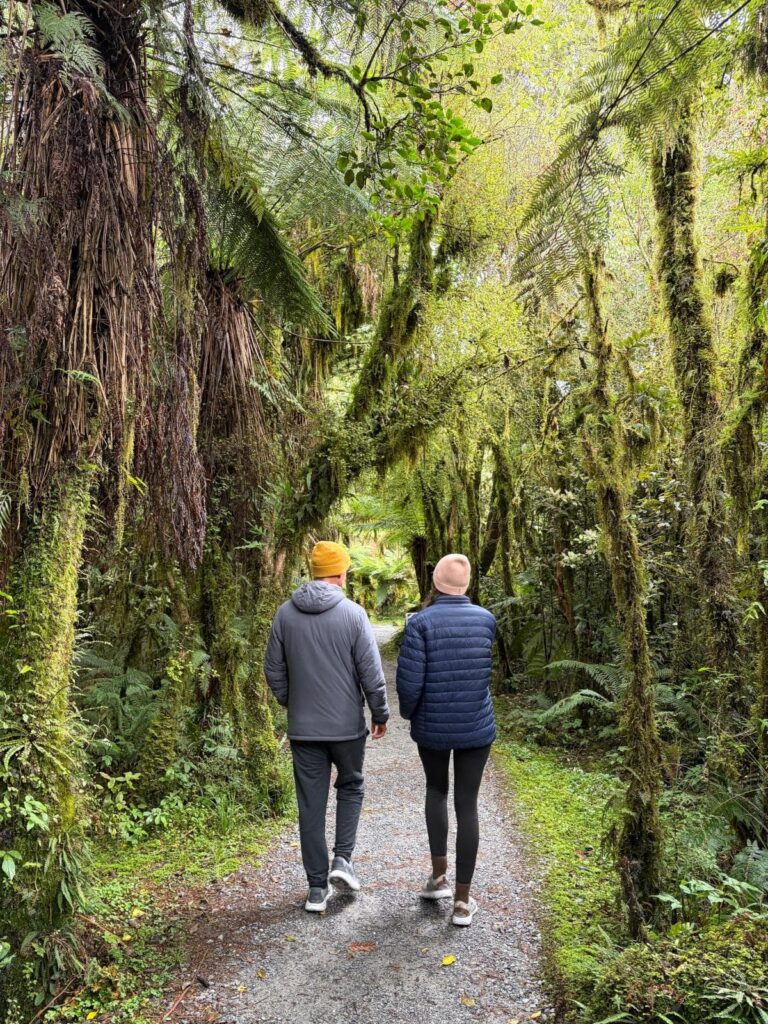 Allan and Katharina walking down a gravel path through a lush, mossy forest on the Haast Pass, New Zealand's West Coast, surrounded by ferns and hanging vines.