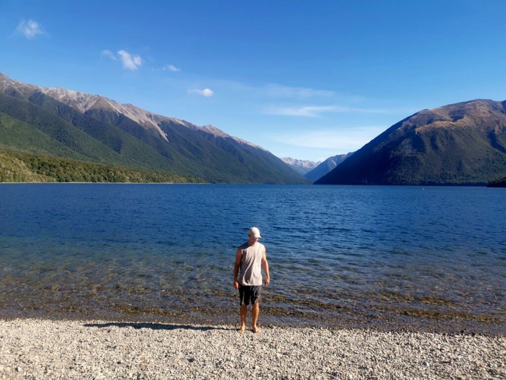 Allan standing on the pebbled shore of Lake Rotoiti, with steep, mountain slopes rising on either side under a clear sky.