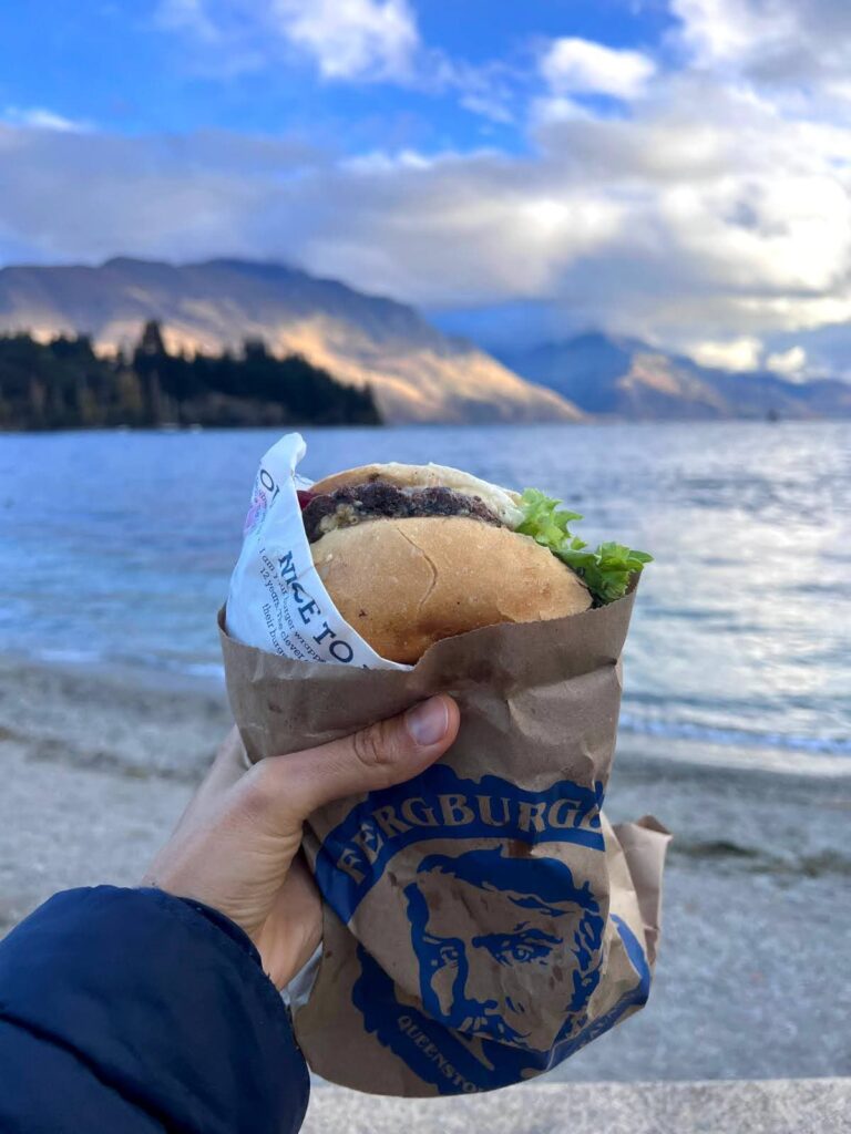 Katharina holding up a large Fergburger in a brown paper bag in Queenstown with Lake Wakatipu and the mountains in the background.