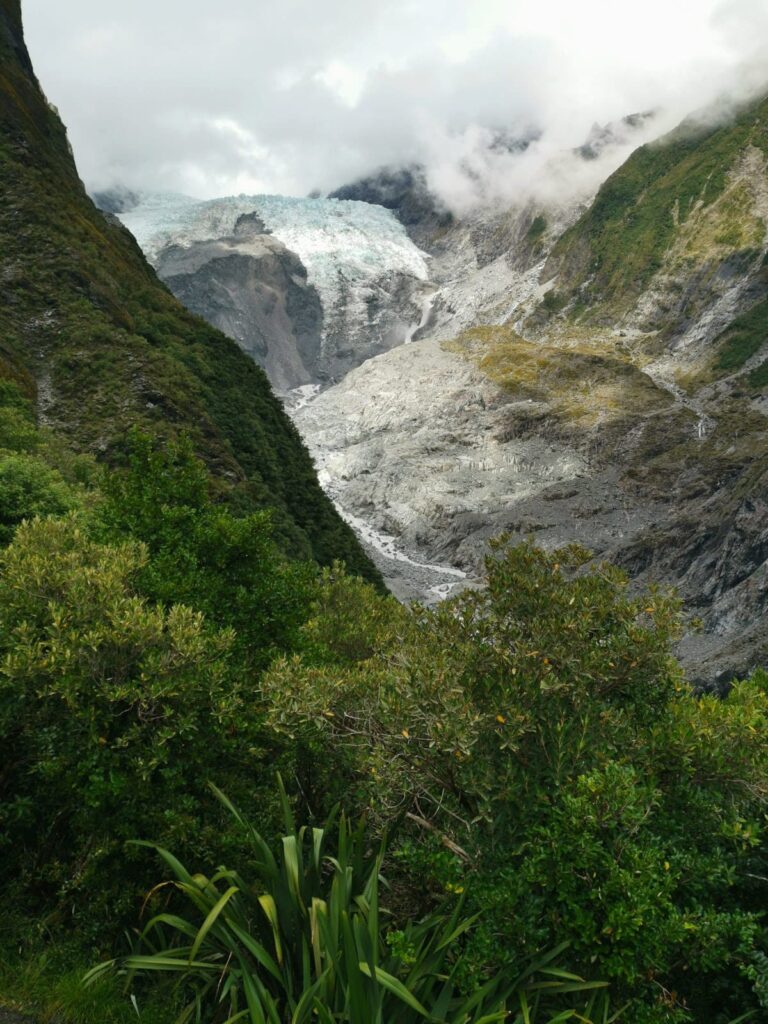 View of Franz Josef Glacier from the Roberts Point Track view point. A light blue and white ice cap sitting on a rocky valley surrounded by native, green plants.