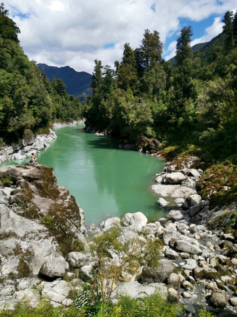 A woman sitting on a rock overlooking the blue-green waters of the Hokitika Gorge winding through rocky banks and lush native forest under a blue sky with white clouds.