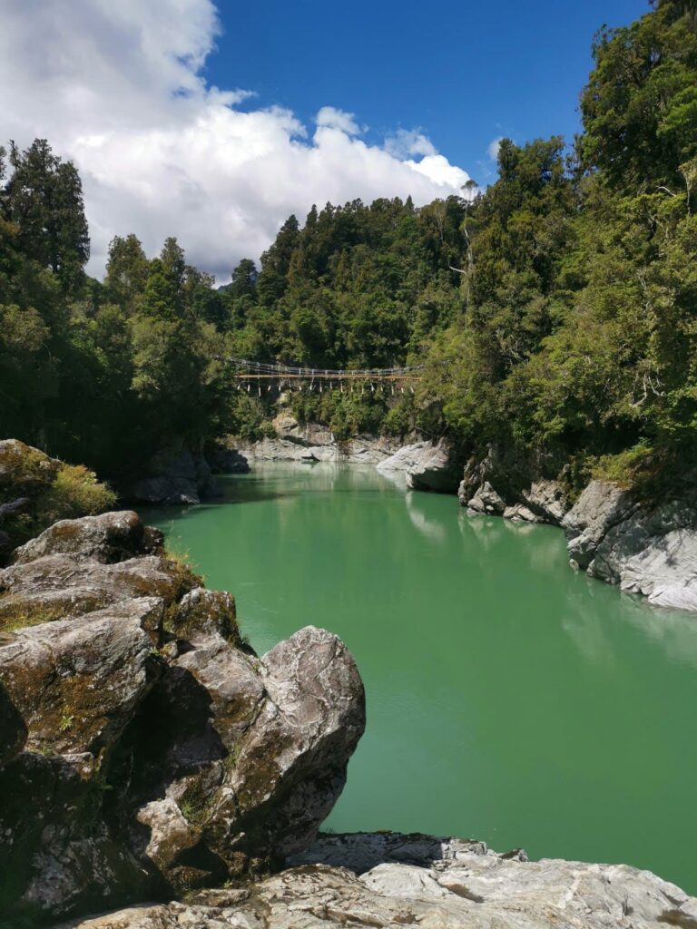 A long brown swing bridge over the blue-green water of Hokitika Gorge, framed by rocky cliffs and dense native forest.