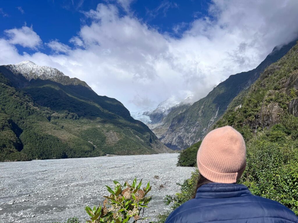 Katharina in a pink beanie looking out over a wide grey glacial riverbed toward the Franz Josef Glacier nestled between green mountains.