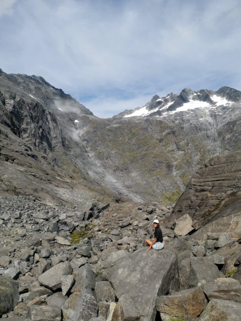 Katharina sitting on a large boulder in a massive field of grey rocks at Gertrudde Saddle with towering, snow-dusted mountains in the background.