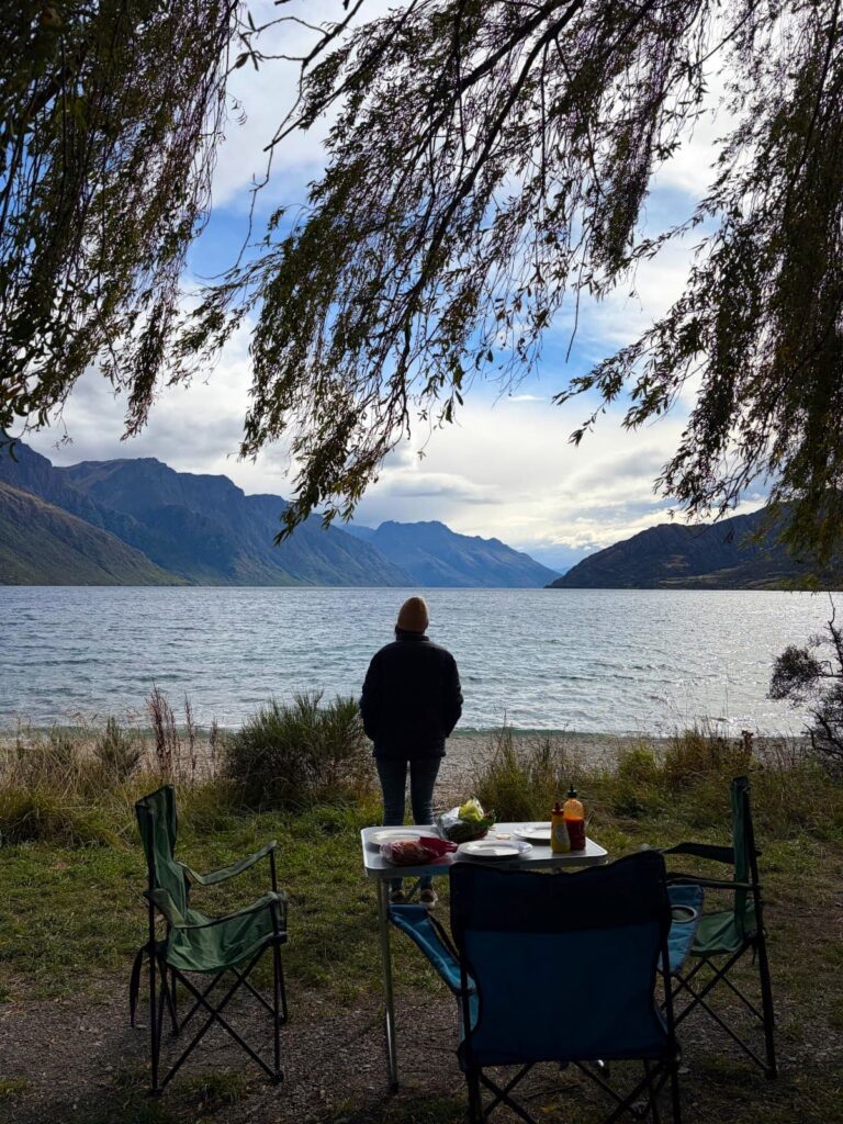 Katharina standing by a camping table set for dinner, looking out at a wide lake and mountains under a willow tree at Kingston Lake Camp.